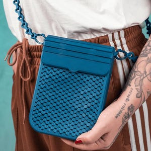 May include: A teal crossbody bag with a geometric pattern and a matching chain strap. The bag has a rectangular shape with a flap closure. The bag is held by a person wearing a white shirt and brown pants.