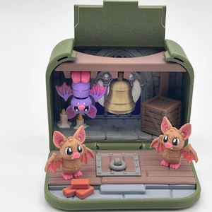 May include: A miniature olive green diorama case featuring a stone-like interior with a hanging purple bat, a gold-colored bell, a brown wooden crate, and two standing tan bats with pink wings. The floor has a central metal ring and red bricks.