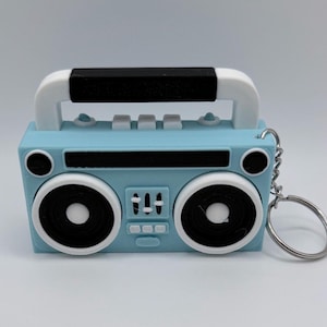 May include: A light blue boombox keyring with white and black details. The keyring has a handle, speakers, and buttons. The speakers are black with white centres. A silver key ring is attached.