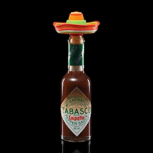 May include: A bottle of Tabasco Chipotle Pepper Sauce with a miniature sombrero bottle stopper. The bottle has a green and white label with the brand name and product information. The sombrero is orange, green, and yellow.