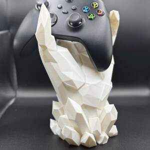 May include: A white, geometric-shaped controller stand, resembling a hand holding a black Xbox controller. The stand has a crystal-like design and is set against a dark background. The controller features colored buttons.