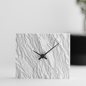 May include: A square white clock with a textured surface. The clock has black hands and a black center. The background is white with a blurred black pot and plant.