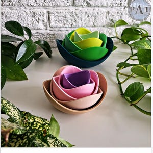 May include: Two sets of nesting bowls in a flower shape. One set features shades of green and yellow, while the other has pink, purple, and tan hues. The bowls are arranged on a white surface with green plants in the background.