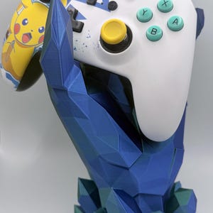 May include: A white video game controller with yellow thumbsticks and a Pikachu graphic on the left grip, held by a blue, low-poly, geometric crystal-shaped stand with dark green accents at the base, featuring A, B, X, and Y action buttons.