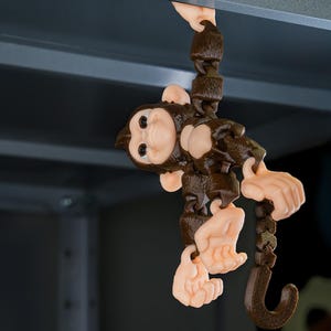 May include: A brown monkey toy with a hook on its tail, hanging from a metal surface. The monkey has a brown body and a light brown face with a playful expression.