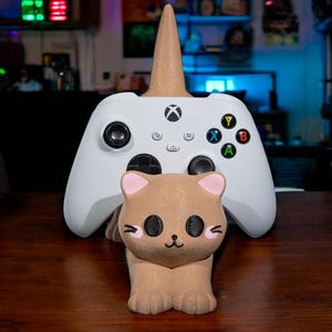 以下が含まれることがあります： 茶色のユニコーンの角が付いた白いXboxコントローラー。ピンクの耳と黒い目の茶色の猫の置物がコントローラーの前にあります。コントローラーには黒いボタンと色の付いたアクションボタンがあります。