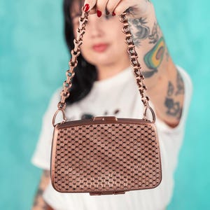 Puede incluir: Un bolso de color cobre con una correa de cadena. El bolso tiene un patrón tejido en la parte delantera y un cierre de broche. La correa de cadena está hecha de eslabones entrelazados. El bolso es de estilo vintage.