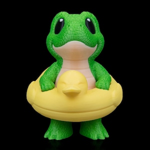 May include: A green frog figurine with large black eyes and a smiling expression. The frog is wearing a yellow duck-shaped floatie. The figurine is set against a black background.