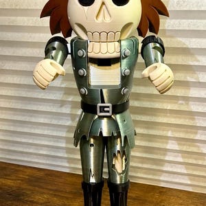 May include: A nutcracker figure with a skull-like head, brown hair, and a metallic green uniform. The figure wears a top hat, has black boots, and stands on a circular base next to a small orange jack-o'-lantern with a green stem.
