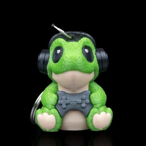 May include: A green frog figurine wearing black headphones and a gray controller. The frog has large black eyes and light tan feet and belly. A silver keychain is attached to the figurine.