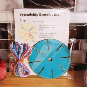 May include: Friendship bracelet kit with colorful string and a blue plastic loom with numbered slots. The instructions say to "Stick the knot through the center hole of the wheel." and "Leaving slot #1 open, put one string into each of the remaining 7 slots."