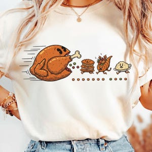 May include: Cream-colored t-shirt featuring a cartoon Thanksgiving theme. A running roasted turkey with a bone chases after a pie, a cornucopia, and mashed potatoes. The design includes colorful dots and a whimsical style.