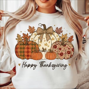 Op de afbeelding: Witte sweatshirt met een Thanksgiving-ontwerp. De afbeelding toont een grote pompoen met een strik, kleinere pompoenen en herfstbladeren. De woorden "Happy Thanksgiving" zijn in script geschreven.