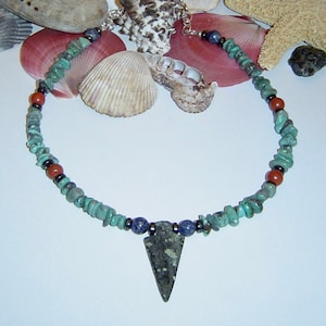 May include: A turquoise and red beaded necklace with a triangular pendant made of dark green stone. The necklace is displayed on a white surface with seashells.