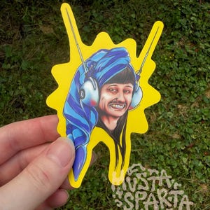 May include: A die-cut sticker featuring a cartoon illustration of a person with a blue striped towel wrapped around their head, wearing white headphones with antennas, and smiling with gold teeth, set against a bright yellow background.