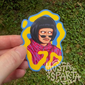 May include: A die-cut sticker featuring a character with a black bob haircut, a thin moustache, and white glowing eyes wearing dark sunglasses. The character wears a red and blue patterned scarf against a blue background with yellow organic shapes.