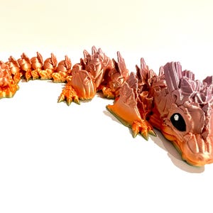 May include: A 3D printed dragon figurine with a gradient color scheme of orange, pink, and purple. The dragon is curled up in a resting pose with its head resting on its tail.
