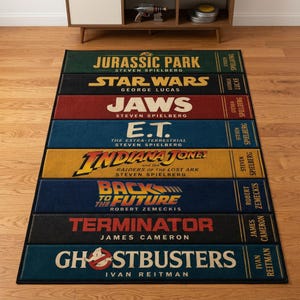 May include: A rectangular rug featuring movie titles like "Jurassic Park," "Star Wars," and "Jaws." Each title is displayed on a colored rectangle with the director's name. The rug has a black border and is placed on a wooden floor.