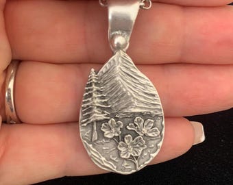 Puede incluir: Un collar con colgante de plata con una escena de la naturaleza. El colgante, con forma de lágrima, presenta una montaña, un árbol y flores. La cadena del collar también es de plata.