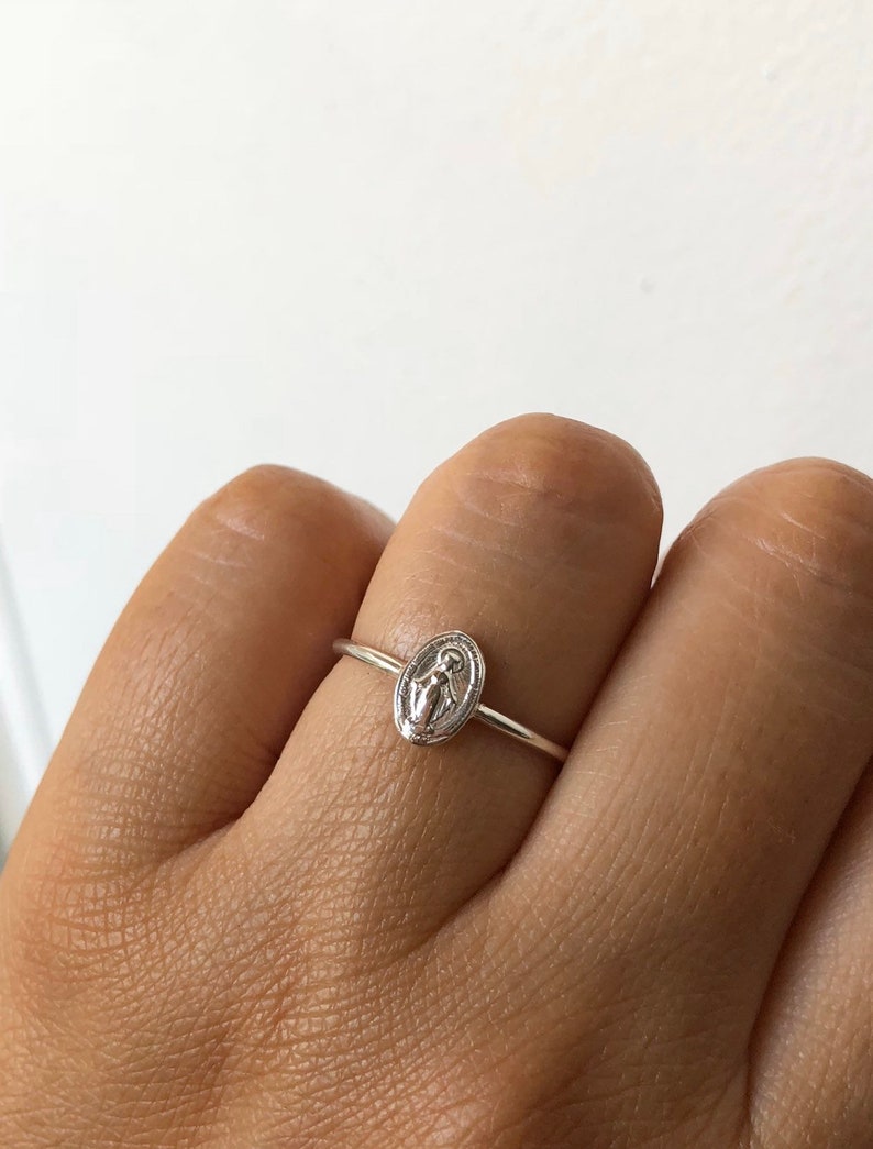 May include: A silver ring with a small oval-shaped charm featuring a depiction of the Virgin Mary.
