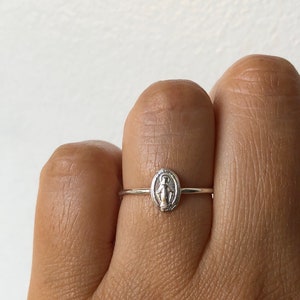 May include: A silver ring with a small oval-shaped charm featuring a depiction of the Virgin Mary.