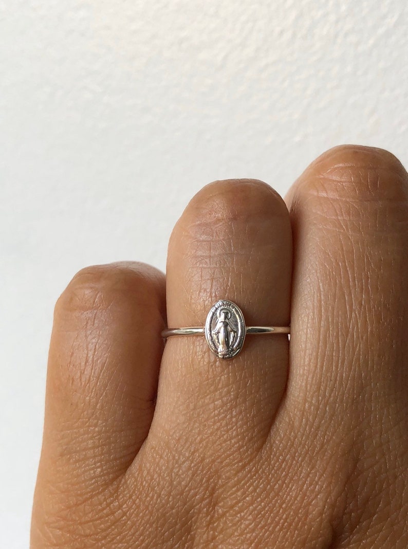 May include: A silver ring with a small oval-shaped charm featuring a depiction of the Virgin Mary.