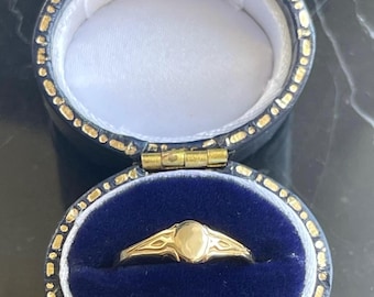 May include: A gold ring with an oval center stone, set in a decorative band. The ring is displayed in an open, blue velvet lined jewelry box with gold trim. The box is a dark blue color.