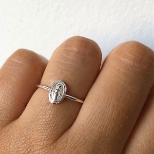 May include: A silver ring with a small oval-shaped charm featuring a depiction of the Virgin Mary.
