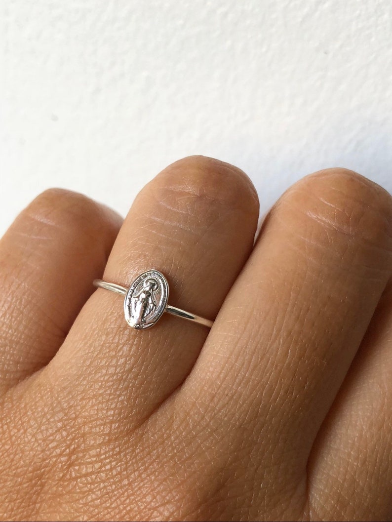 May include: A silver ring with a small oval-shaped charm featuring a depiction of the Virgin Mary.