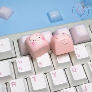 May include: Close-up of a keyboard with custom keycaps. Two pink keycaps shaped like pigs are the focal point, with white keys and a light blue background. The keycaps have pig faces with blush and small details.
