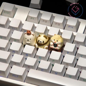 May include: Three small animal figurines, a white polar bear, a brown bear with a rope belt, and a hamster holding a small black object, are placed on a white keyboard with black keys.