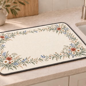 May include: A rectangular kitchen drying mat with a cream-colored base and a dark gray border. The mat features a printed botanical pattern of wildflowers, including red, white, and blue blossoms with green foliage arranged along the edges.