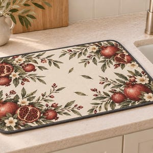 May include: A rectangular dish drying mat featuring a cream-coloured background with a botanical print of red pomegranates, green leaves, and small white flowers. The mat has rounded corners and a dark grey fabric border trim around the entire edge.