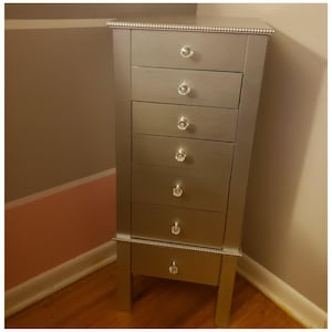 May include: A silver-colored jewelry armoire with six drawers and a bottom cabinet. Each drawer has a crystal-like knob. The armoire is tall and narrow, with a beaded trim around the top and bottom.