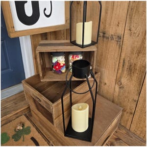 May include: A rustic home decor arrangement featuring wooden crates, black metal lanterns with cream-colored LED candles, and jars filled with colorful decorative items. The scene is set against a backdrop of wooden planks.