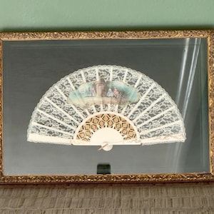 May include: A framed vintage hand fan with a gold ornate frame. The fan features a white lace design with a painted scene at the top. The fan's handle is a cream color with a gold geometric pattern.