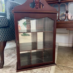 May include: A dark wood display cabinet with a glass door, featuring a decorative arched top with a central finial, a brass-toned metal latch, and multiple interior shelves divided into compartments for organizing and showcasing small items.