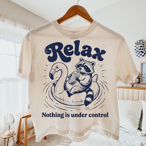 Könnte beinhalten: Cremefarbenes T-Shirt mit einem marineblauen Aufdruck eines Waschbären auf einem Flamingo-Schwimmring, der aus einem Becher trinkt. Das Wort "Relax" steht über der Grafik, darunter "Nothing is under control".