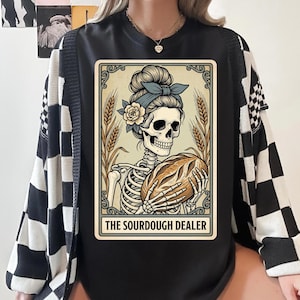 Könnte beinhalten: Schwarzes T-Shirt mit einer Skelett-Illustration, das Brot hält, umrahmt von Weizenähren. Der Text lautet "THE SOURDOUGH DEALER". Die Person trägt eine schwarz-weiß karierte Strickjacke.