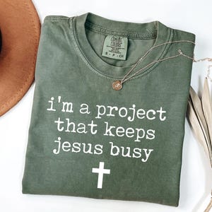 Puede incluir: Camiseta verde oliva con el texto "I'm a project that keeps Jesus busy" y una cruz blanca. Un collar dorado con un colgante descansa sobre la camiseta. Un sombrero marrón y hojas de palma secas están en el fondo.