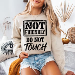 May include: A beige sweatshirt with the text "NOT FRIENDLY DO NOT TOUCH" in a black, distressed font. The text is centered on the front of the long-sleeved top. The sweatshirt is worn with denim shorts.