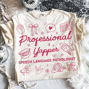 Puede incluir: Camiseta color crema con las palabras "Professional Yapper" en cursiva, junto con el texto "Speech Language Pathologist". La camiseta presenta ilustraciones rojas de un cerebro, un lazo, una burbuja de diálogo, un teclado, una boca, un helado, un megáfono, una mano, una flor, una oreja y un lazo.
