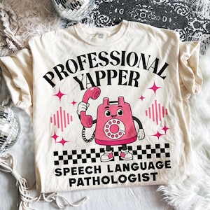 Puede incluir: Camiseta color crema con las palabras negras "PROFESSIONAL YAPPER SPEECH LANGUAGE PATHOLOGIST". Un gráfico de teléfono de dibujos animados rosa está en el centro, con estrellas rosas y acentos de ondas de sonido. La parte inferior tiene un patrón de cuadros blanco y negro.