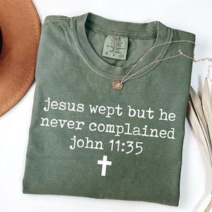 May include: Olive green t-shirt with the text "Jesus wept but he never complained John 11:35" and a cross. A gold necklace with a pendant rests on the shirt. The shirt is folded on a white surface.