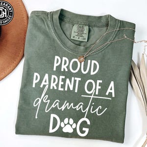 Puede incluir: Camiseta verde oliva con el texto blanco "PROUD PARENT OF A dramatic DOG". El diseño incluye una huella de pata y un collar dorado. La camiseta es de la marca Comfort Colors.