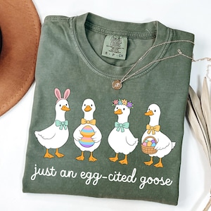 May include: Olive green Comfort Colors t-shirt featuring a graphic of four cartoon geese, each adorned with Easter-themed accessories like bunny ears and flower crowns. The text "just an egg-cited goose" is printed below the geese.