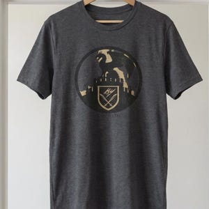 Puede incluir: Camiseta gris oscuro con un dise&ntilde;o circular que muestra un drag&oacute;n sobre un castillo, un escudo y la palabra "RAWNATH" debajo. La camiseta est&aacute; colgada en una percha de madera contra una pared blanca.