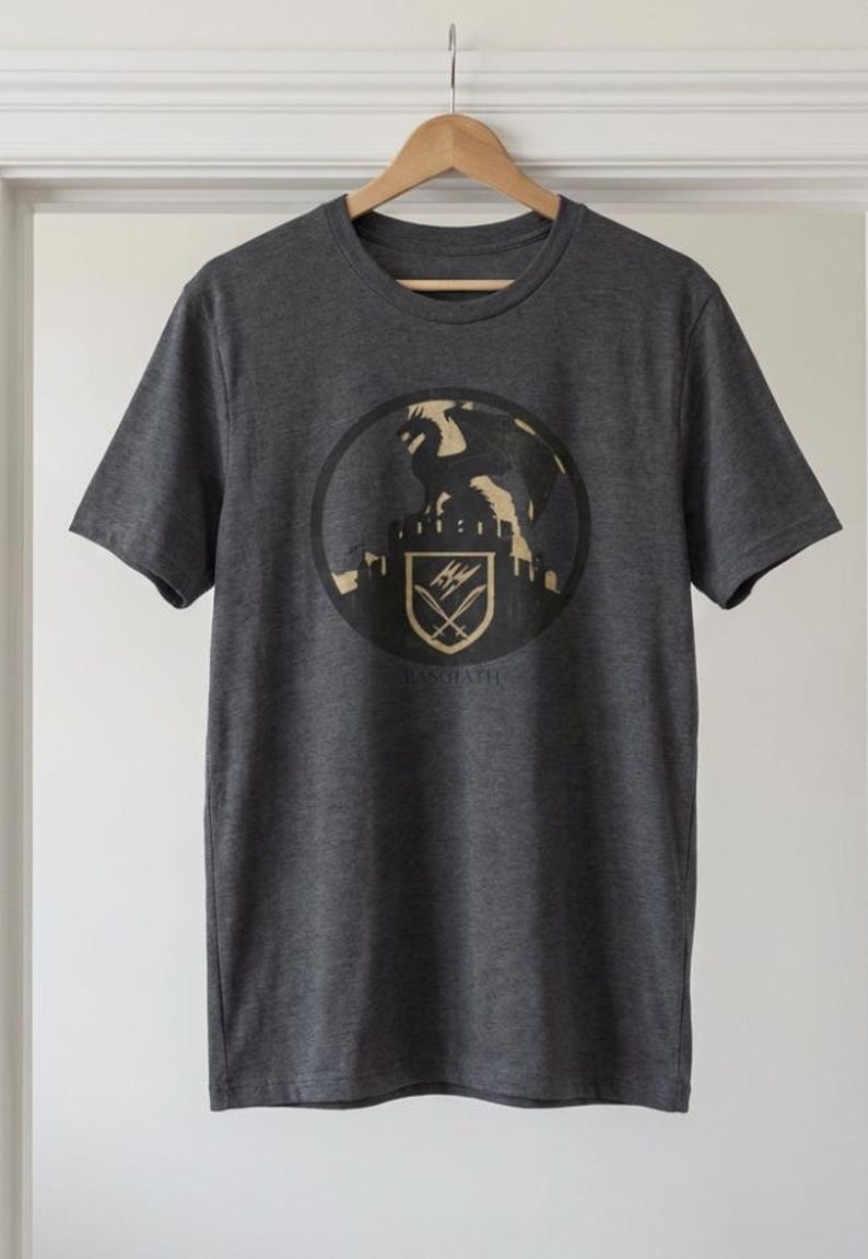 Puede incluir: Camiseta gris oscuro con un dise&ntilde;o circular que muestra un drag&oacute;n sobre un castillo, un escudo y la palabra "RAWNATH" debajo. La camiseta est&aacute; colgada en una percha de madera contra una pared blanca.