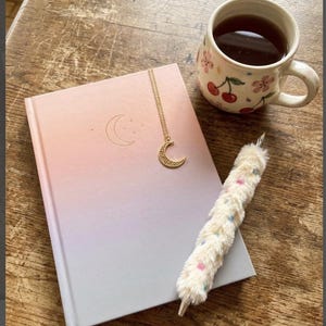 May include: A journal with a gradient pink and gray cover, a gold crescent moon necklace, and a fluffy pen with pastel dots rest on a wooden surface. A floral-decorated mug filled with tea sits nearby.