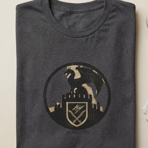 Puede incluir: Camiseta gris oscuro con un dise&ntilde;o circular. El dise&ntilde;o presenta un drag&oacute;n sobre un castillo, un escudo con espadas cruzadas y la palabra "BASGIATH" debajo. El dise&ntilde;o es en negro y beige.